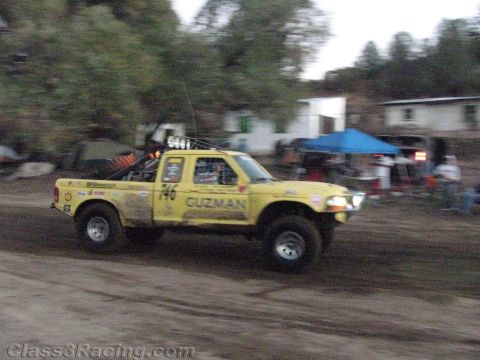 1990 Ford Bronco - yellow off-road race truck numbered 746 and lettered 'Guzman' kicks up dust on a dirt track past spectators and a blue tent.