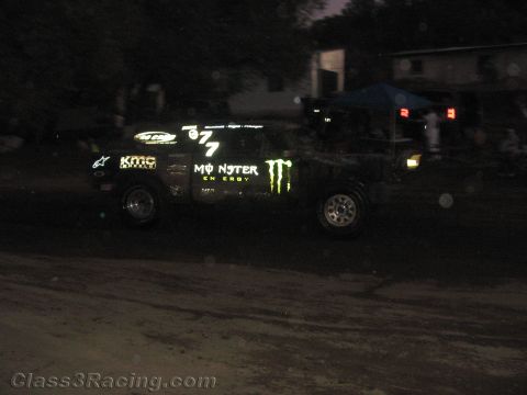 1990 Ford Bronco - dark off-road race truck wearing Monster Energy and KMC livery, numbered 877, races on a dirt course at night.
