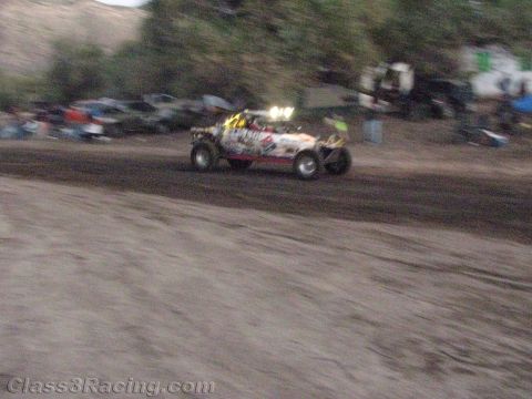 1990 Ford Bronco - open-wheel off-road race buggy with roof lights speeds across a dusty track at dusk past a row of parked vehicles.