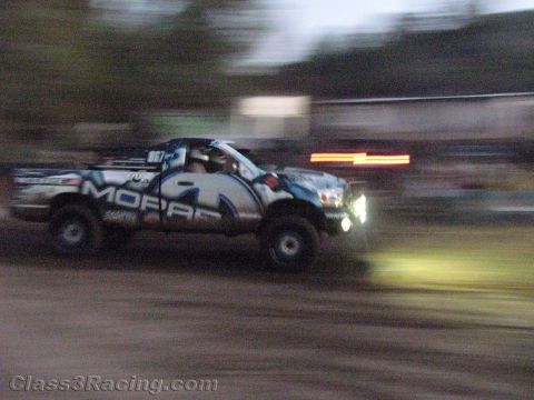 1990 Ford Bronco - blue-and-white off-road race truck lettered 'Mopar' races on a dirt course at dusk with headlights on.