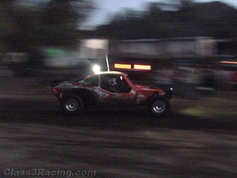 1990 Ford Bronco - orange off-road race buggy with a single roof light blasts along a dirt track at dusk, motion-blurred.
