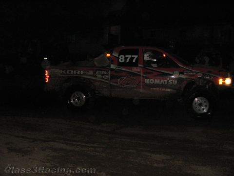 1990 Ford Bronco - dirt-covered off-road race truck numbered 877 with Kore and Komatsu graphics sits on a dark dirt course at night.