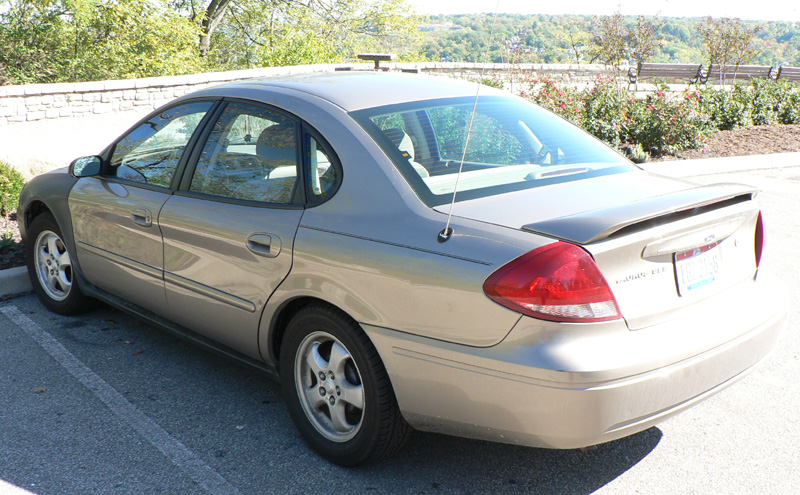 Gold Ford Taurus sedan rear three-quarter view parked in a lot near a stone wall