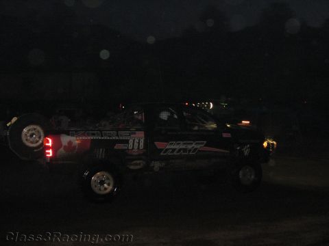 1990 Ford Bronco - dark off-road race truck numbered 868 with Kore and HRT graphics moves along a dirt course at night.