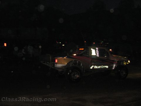 1990 Ford Bronco - off-road race truck bearing 'Mills Customs' lettering sits on a dark dirt course at night amid scattered lights.