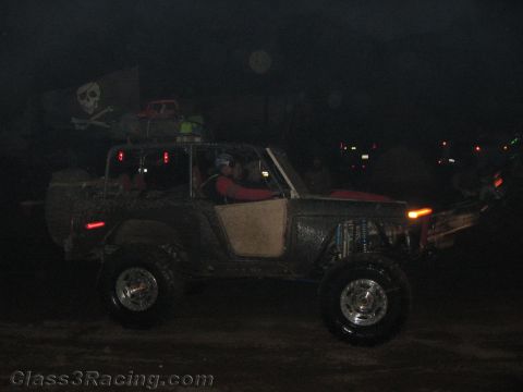 1990 Ford Bronco - muddy open-top off-road buggy flying a Jolly Roger flag and seating a driver in red drives on a dark course at night.