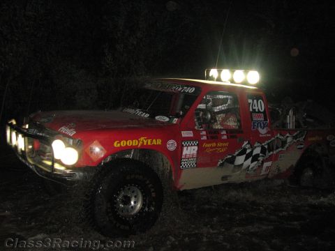 1990 Ford Bronco - red off-road race truck numbered 740 with Goodyear graphics and a roof light bar splashes through a dark course at night.