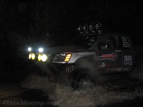 1990 Ford Bronco - silver off-road race truck numbered 1504 with Camburg and Titan graphics throws up spray crossing water at night.