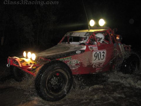 1990 Ford Bronco - red tube-frame off-road race buggy numbered 903 splashes through water with twin headlights blazing at night.