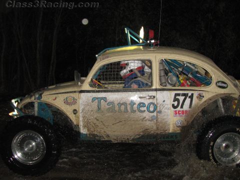 1990 Ford Bronco - muddy beige Volkswagen Beetle Baja race car numbered 571 with 'Tanteo' lettering races on a dark course at night.