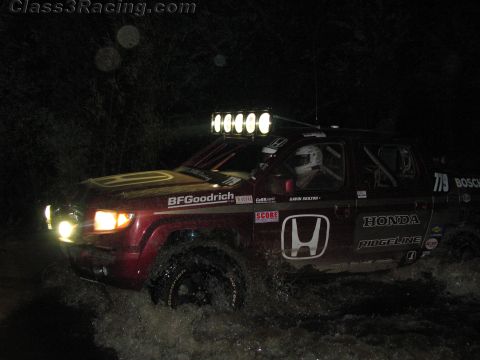 1990 Ford Bronco - dark red Honda Ridgeline off-road race truck numbered 779 with a roof light bar splashes through water at night.