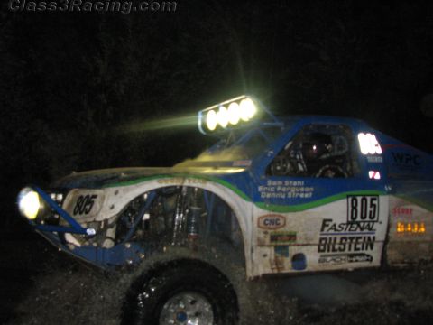 1990 Ford Bronco - blue-and-white off-road race truck numbered 805 with Fastenal and Bilstein graphics races through a dark course at night.