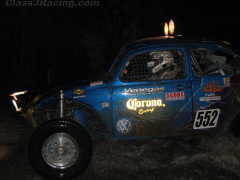 1990 Ford Bronco - blue Volkswagen Beetle Baja race car numbered 552 with Corona and Venegas graphics sits on a dark course at night.