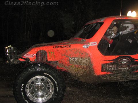 1990 Ford Bronco - muddy orange off-road race buggy with Monster Energy, KMC and Lowrance graphics rolls along a dark course at night.