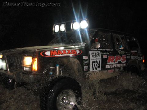 1990 Ford Bronco - Jeep Cherokee off-road racer numbered 1734 with Maxxis and RPS Suspension graphics splashes through water at night.