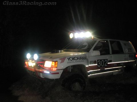 1990 Ford Bronco - white Jeep Grand Cherokee race truck numbered 302 with a glowing roof light bar churns through mud at night.