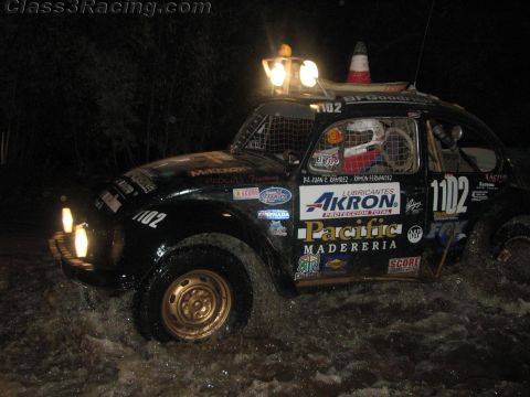 1990 Ford Bronco - black Volkswagen Beetle Baja race car numbered 1102, covered in sponsor decals, throws spray crossing water at night.