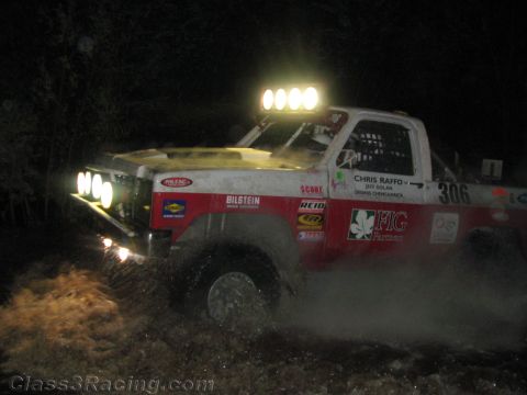 1990 Ford Bronco - white and red race pickup numbered 306 with a roof light bar sprays mud and water while racing at night.