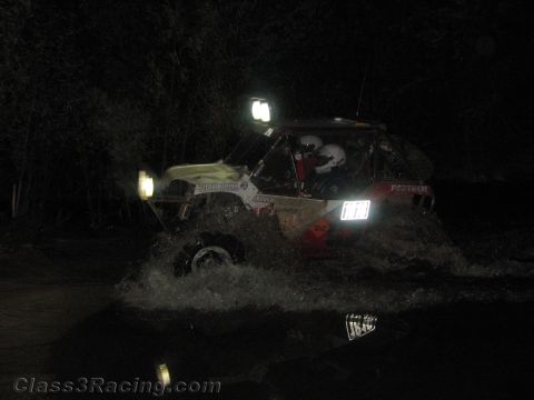 1990 Ford Bronco - pale off-road race vehicle crosses a water section at night, headlights blazing as it throws up a sheet of spray.
