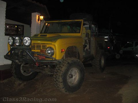 1990 Ford Bronco - yellow lifted early Ford Bronco with triple roof lights and big tires is parked beside a stucco building at night.