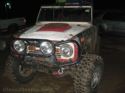 1990 Ford Bronco - muddy front end of a weathered white early Ford Bronco prerunner with round headlights and a red hood scoop at night.