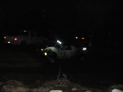 1990 Ford Bronco - dimly lit off-road race vehicle with roof lights and number plate sits beside a white pickup in a dark staging area.