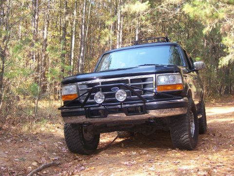 1996 Ford Bronco - A black Ford Bronco with off-road modifications is parked on a dirt path surrounded by trees.