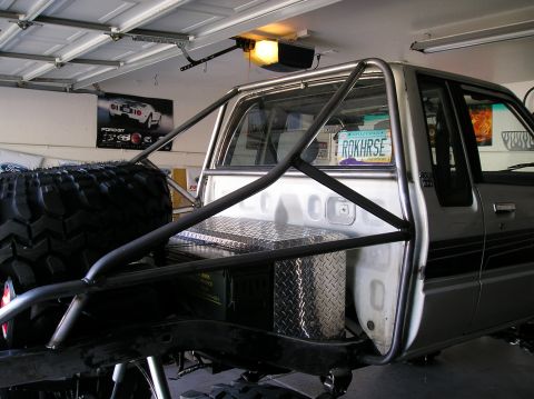 1986 Toyota Hilux - Bare-steel exo cage wrapping the bed and cab of a white Toyota pickup in a garage, with a diamond-plate toolbox and green ammo can in the b…