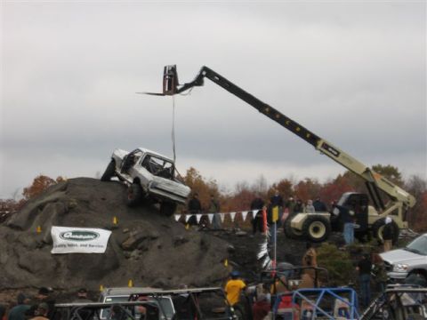 1995 Ford Bronco - A white pickup truck is lifted by a telehandler during an off-road event, with spectators gathered below.