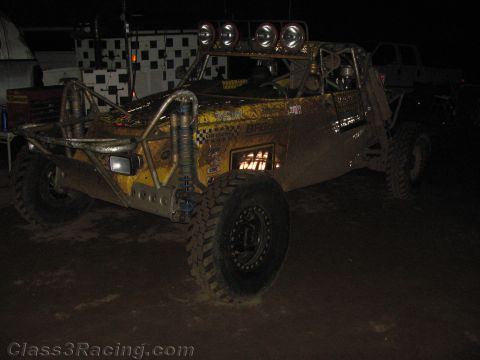 1990 Ford Bronco - mud-caked yellow tube-frame off-road buggy with roof lights sits in front of a checkered banner at night.