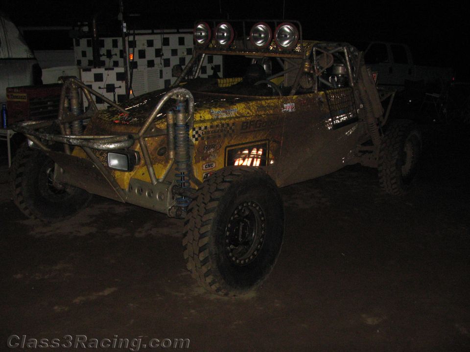 Muddy yellow tube-frame off-road buggy in front of checkered banner at night