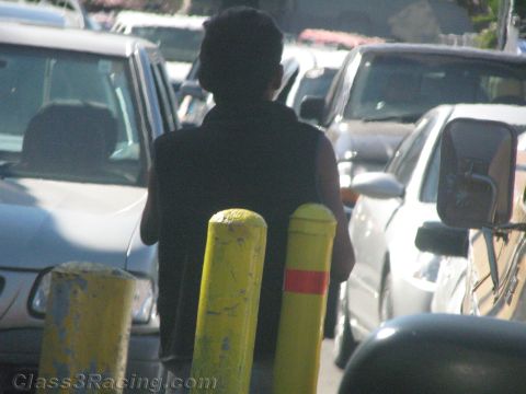1990 Ford Bronco - person in a black tank top walks away between rows of parked silver cars, passing two yellow safety bollards.
