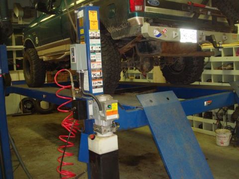 1996 Ford Bronco - A green Ford truck with large off-road mud tires raised on a blue four-post hydraulic lift inside a garage, showing the undercarriage, soli…