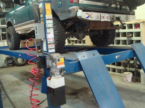 1996 Ford Bronco - A dark green Ford Bronco is raised on a blue drive-on lift in a garage, viewed from the front-left underside showing large off-road tires,…