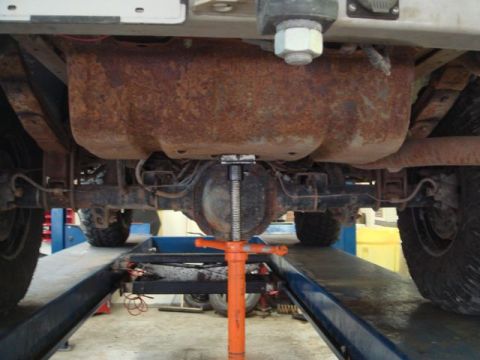 1996 Ford Bronco - Underbody view of a vehicle raised on a lift, showing a heavily rusted fuel tank or differential housing with an orange floor jack supporti…