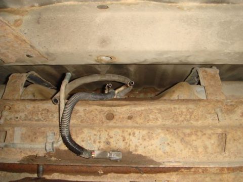 1996 Ford Bronco - Underbody view of a rusty vehicle chassis showing a fuel tank area with two cut/open metal fuel or vent lines, a corrugated wire harness co…