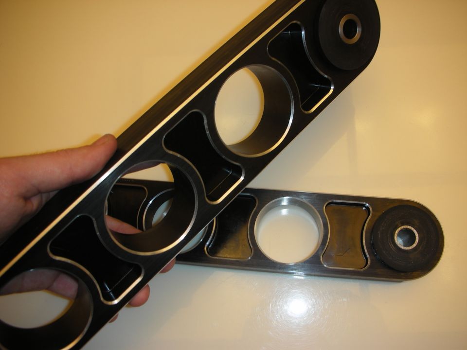 Hand holding a black machined aluminum control arm beside another arm