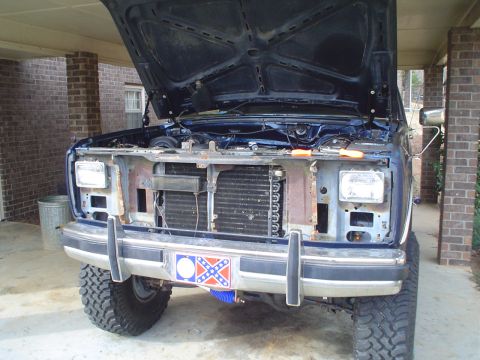 1984 Ford Bronco - Front view of a dark blue SUV with its hood open, showing missing grille and headlights. A rebel flag decal is visible on the bumper.