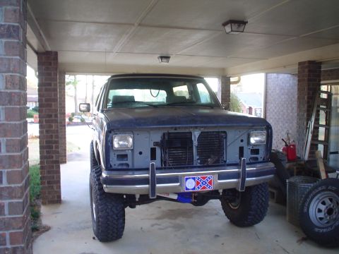 1984 Ford Bronco - This dark blue SUV is parked under a carport, showing its front end with a missing grille and headlights, and lifted suspension.