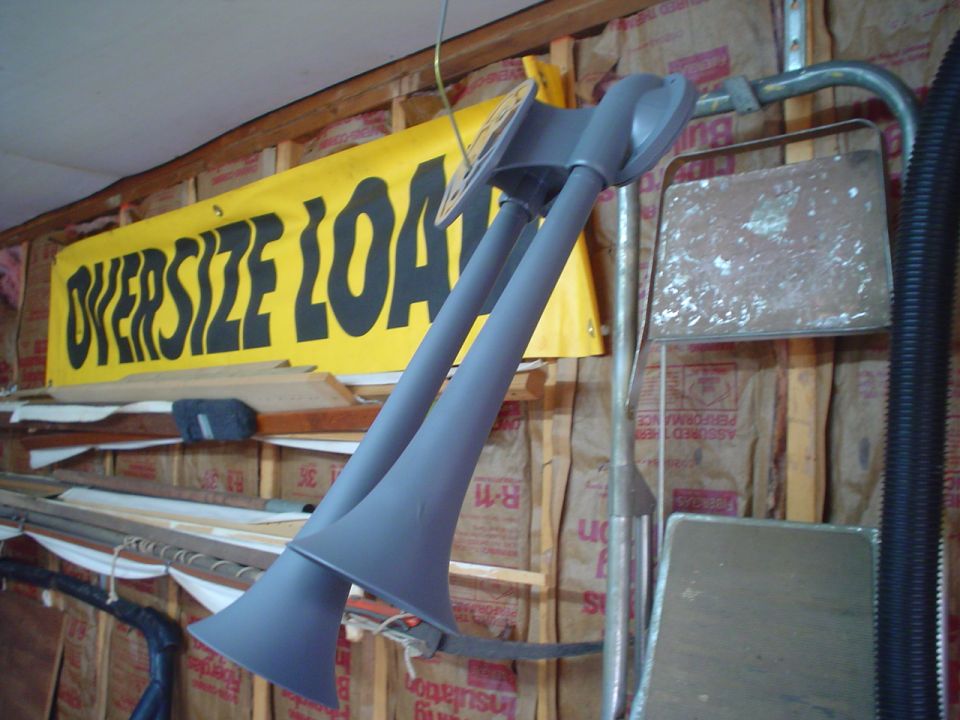 Pair of gray air horns hanging in a garage, yellow 'OVERSIZE LOAD' banner in background