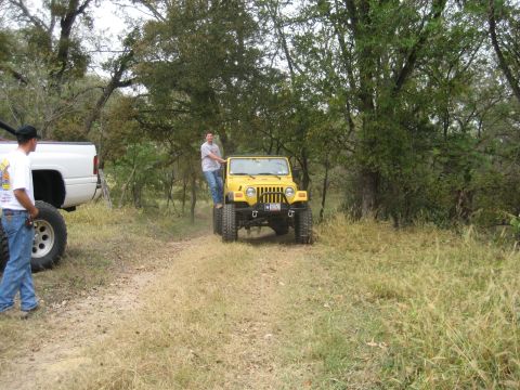 2001 Jeep Wrangler - Two vehicles, a yellow Jeep Wrangler and a white pickup truck, are on a dirt path surrounded by trees.