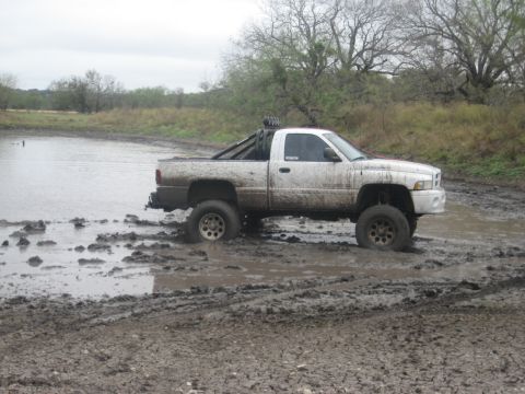 2001 Jeep Wrangler - A white pickup truck is stuck in deep mud, with trees and water in the background.