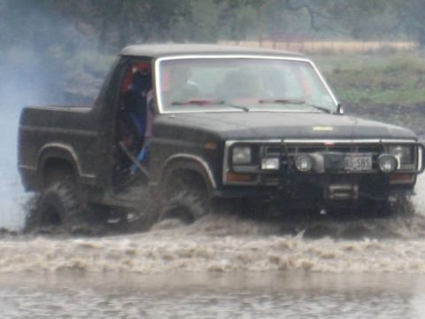 2001 Jeep Wrangler - A black Ford Bronco II navigates through murky water, kicking up a spray. The vehicle appears to be from the 1980s.