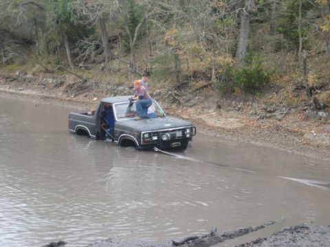 2001 Jeep Wrangler - A gray pickup truck is stuck in muddy water, with two individuals perched on its hood as a tow rope is attached.