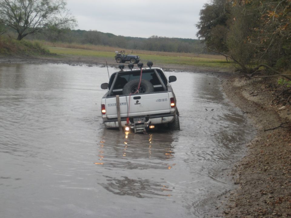 White pickup truck with spare tire in bed driving through muddy water; another off-road vehicle in background