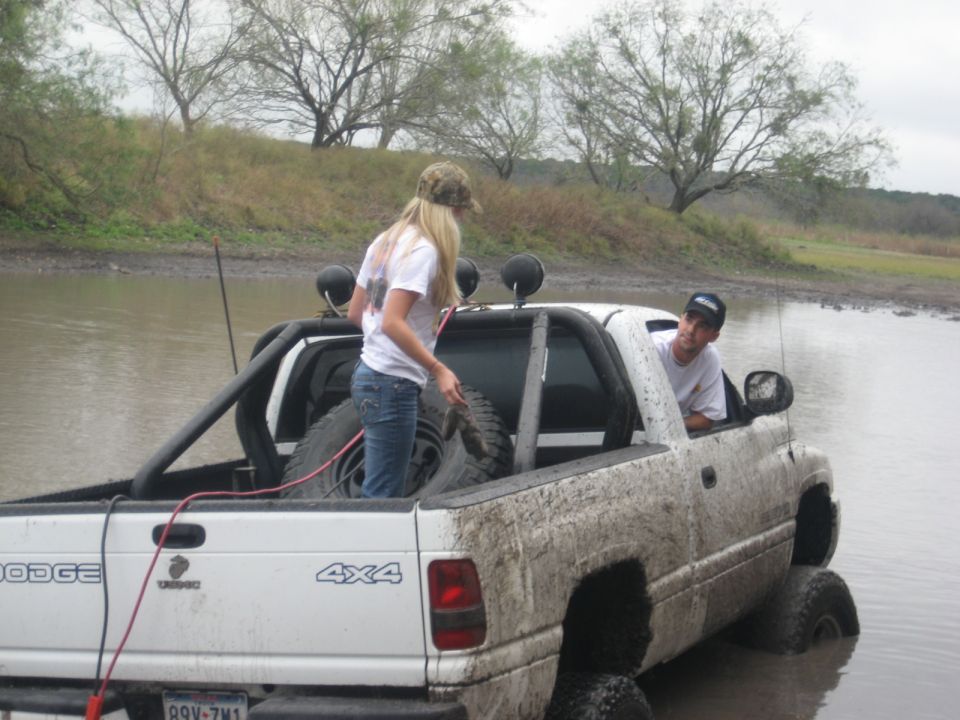 White Dodge Ram 4x4 stuck in muddy water, woman in bed, man in driver seat