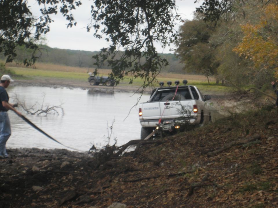 White pickup truck stuck in muddy water, person pulling a strap from the side. ATV in background