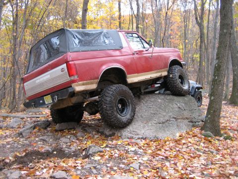 1990 Ford Bronco - A red Ford Bronco navigates a rocky trail during autumn, with another vehicle visible behind it.