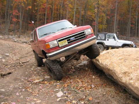 1990 Ford Bronco - Red 1990 Ford Bronco navigating an obstacle on an off-road trail during autumn.