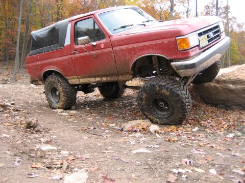 1990 Ford Bronco - A red 1990 Ford Bronco is shown off-roading on a rocky trail with autumn foliage in the background.